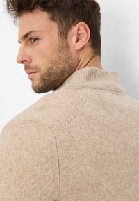 Beiger Strickpullover mit hohem Kragen und strukturiertem Stoff, mit Raglanärmeln und sichtbaren Nahtdetails.
