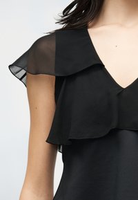 Γυναίκα που φοράει μαύρο αμάνικο τοπ με διαφανές ριζάτο επάνω στρώμα και V-λαιμόκοψη, εμφανίζοντας τον ώμο και το πάνω μέρος του στήθους.