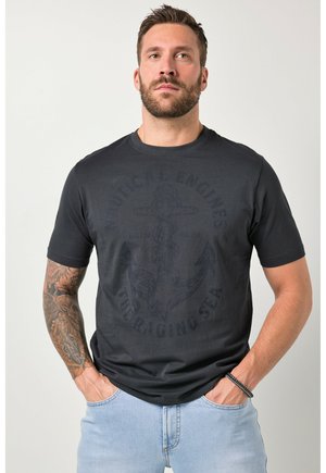 Man med skägg och tatuerad högra arm bär en mörk t-shirt med ankartema och ljusblå jeans, händerna i fickorna, mot en enkel vit bakgrund.