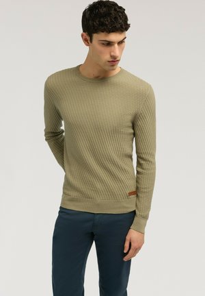 Joven con suéter verde oliva texturizado y pantalones azul oscuro, de pie con los brazos detrás de la espalda, mirando hacia abajo y de lado.