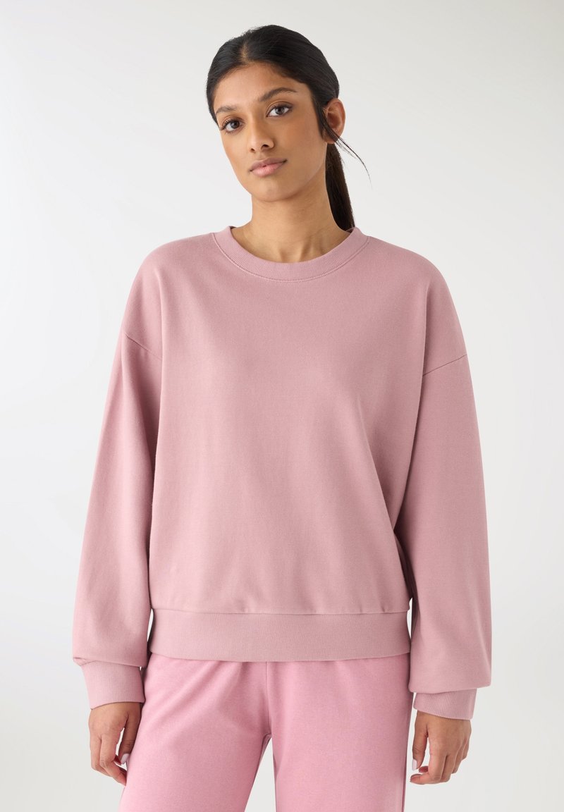 Felpa oversize rosa chiaro realizzata in materiale morbido, con scollo rotondo e polsini a costine. Abbinata a pantaloni coordinati.