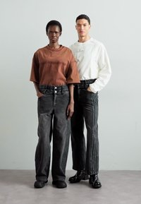 Model wearing an oversized brown shirt and black baggy jeans with vertical seams. Another model in a white shirt and similar black jeans.