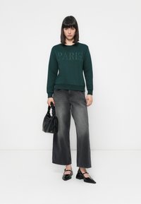 Donna che indossa una felpa verde scuro con la scritta "PARIGI", jeans neri a gamba larga scoloriti, scarpe Mary Jane nere, mentre tiene una borsa nera arricciata.