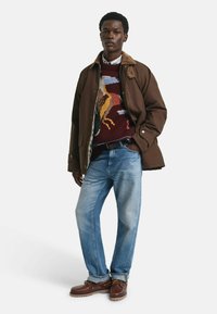 Braune gesteppten Jacke mit Cordkragen, gemustertem Strickpullover in Mahagonifarben und Beige, hellblauen Jeans und braunen Schnürschuhen.