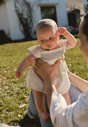 Adulte tenant un bébé en robe blanche à l'extérieur sur l'herbe, bébé touchant l'oreille et regardant vers le bas avec une expression neutre.