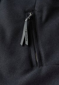 Schwarzer Fleece-Stoff mit einer Reißverschlusstasche. Der Reißverschluss hat einen markierten Ziehschutz. Die Oberfläche hat eine weiche Textur und minimal sichtbare Nähte.