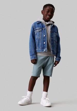Giacca di jeans con bottoni, felpa grigia sotto, pantaloni corti azzurri e sneakers bianche con calzini a coste. Outfit casual per attività sportive.