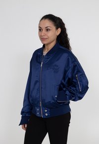 Marineblaue, glänzende Bomberjacke mit gerippten Bündchen und Saum, Reißverschlusstaschen und gesticktem Logo-Detail auf der Vorderseite.