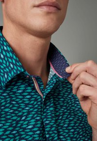 Camicia turchese con motivo di pesci, dotata di un colletto viola a contrasto e dettagli rosa. Il materiale sembra liscio con una vestibilità strutturata.