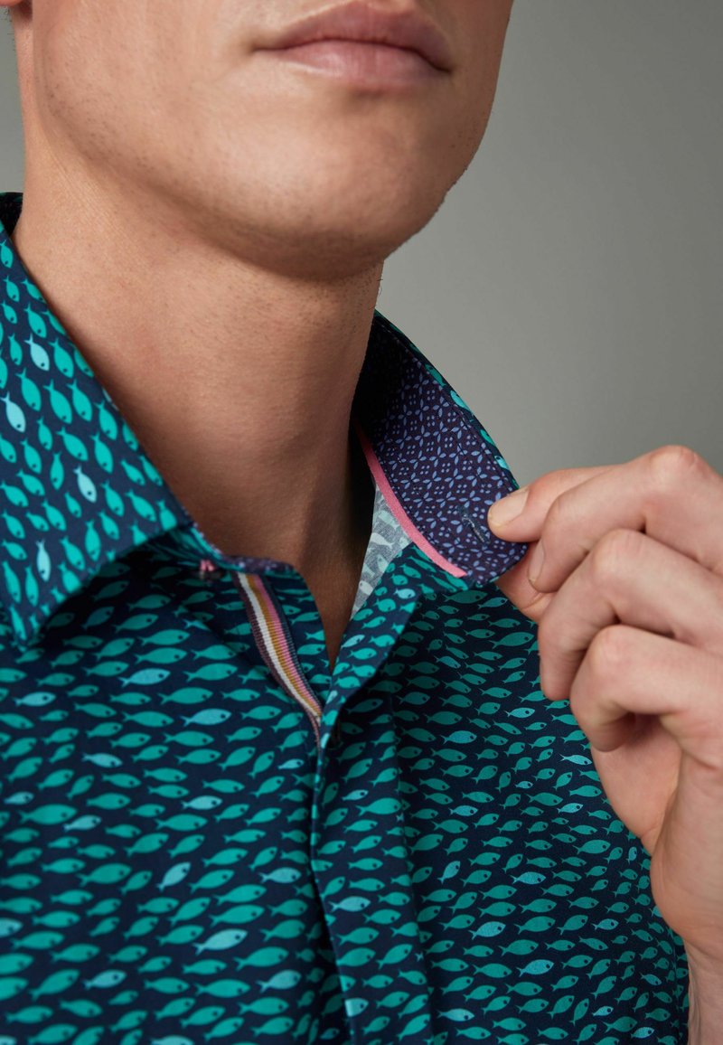 Camicia turchese con motivo di pesci, dotata di un colletto viola a contrasto e dettagli rosa. Il materiale sembra liscio con una vestibilità strutturata.