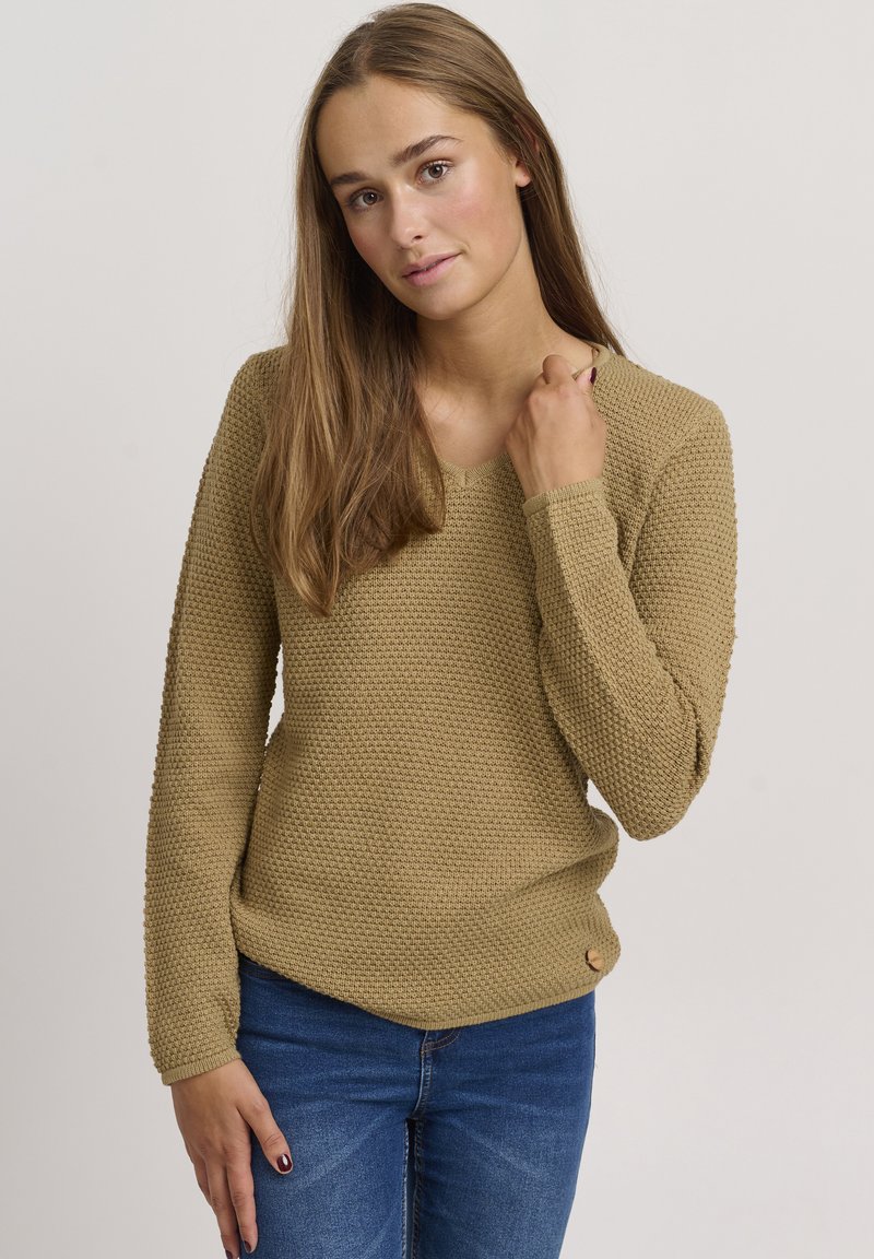 Khaki sweater with a textured, knitted design, V-neckline, long sleeves, and a rounded hem. Paired with blue jeans; a button detail at the side.
