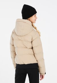 Beige Pufferjacke mit Kapuze, ausgestattet mit einem gerippten schwarzen Saum und einer glatten, leicht glänzenden Textur. Die schwarze Strickmütze setzt einen guten Kontrast.