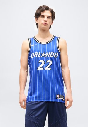 Junger Mann, der ein blaues Orlando-Basketballtrikot mit der Nummer 22 und passende marineblaue Sportshorts trägt, steht vor einem schlichten Hintergrund.