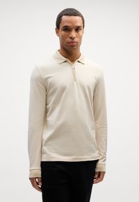 Beige langermet polo skjorte med glidelås, teksturert stoff og klassisk krage. Har kontrastfargede mansjetter og en tettsittende design.