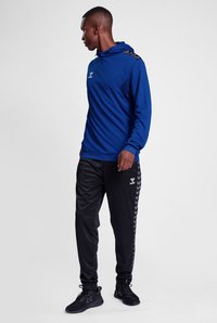 Felpa con cappuccio a maniche lunghe blu con dettagli neri, abbinata a pantaloni sportivi neri con un motivo a zigzag lungo i lati.