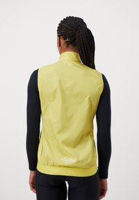 Gelbes, leichtes Radfahrweste mit hohem Kragen, elastischem Saum und reflektierenden Akzenten, getragen über einem schwarzen Langarmoberteil. Branding auf der Rückseite.