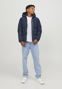 Marinblå puffjacka med huva, med quiltat mönster och en logotyp fram. Bärs över en vit skjorta, ihop med ljusblå jeans.