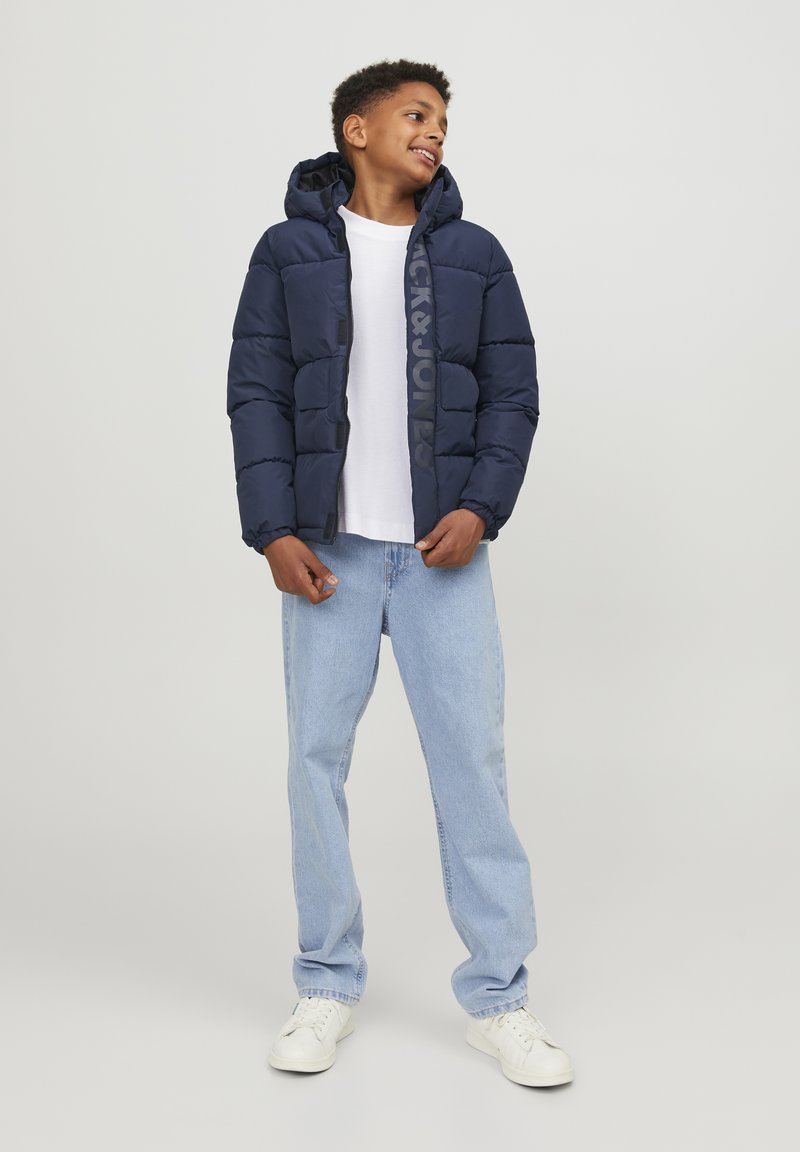 Marinblå puffjacka med huva, med quiltat mönster och en logotyp fram. Bärs över en vit skjorta, ihop med ljusblå jeans.