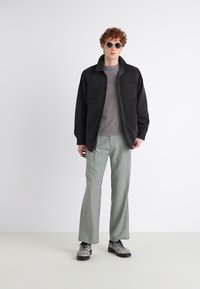 Zwarte spijkerjacket met twee borstzakken over een grijze shirt, gecombineerd met lichtgroene broeken en grijze schoenen. Ronde zonnebril maakt de look compleet.