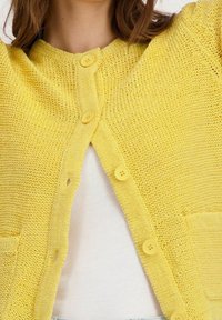 Gul stickad cardigan med rund halsringning, fyra knappar och två framfickor. Strukturerad tyg med en lös passform.