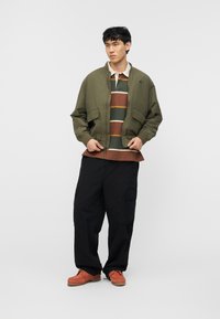 Νεαρός άνδρας που στέκεται φορώντας μια χακί bomber μπουφάν, ριγέ πόλο πουκάμισο, φαρδιά μαύρη cargo παντελόνια και καφέ δερμάτινα παπούτσια.