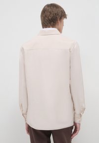 Camicia a maniche lunghe color beige chiaro con vestibilità rilassata, texture sottile, colletto e orlo dritto. Include polsini con bottone e spacco posteriore.