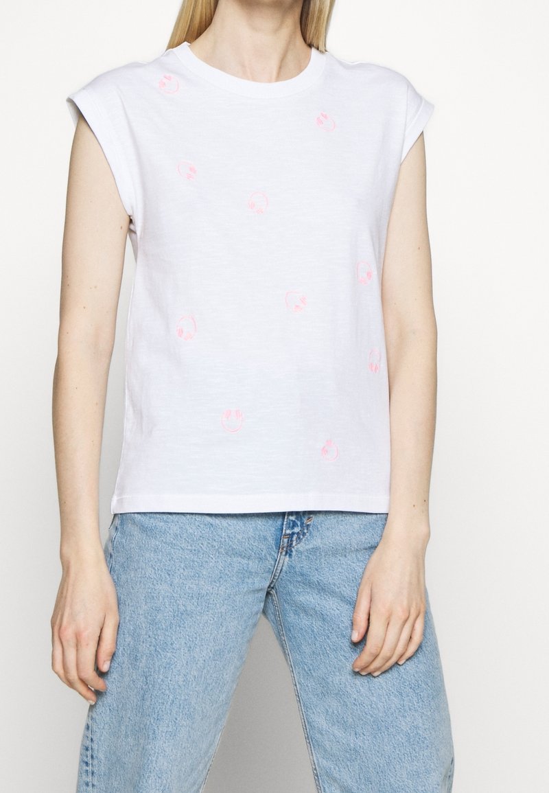 Mujer vistiendo una camiseta sin mangas blanca con pequeños estampados de caras sonrientes rosas y jeans de tiro alto azul claro, contra un fondo blanco.