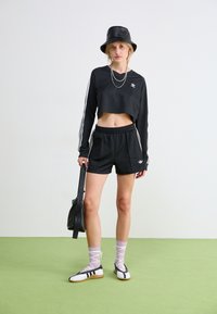 Svart cropped langermet topp med hvite striper, matchende svarte shorts, hvite og svarte sko, og en svart bøttehatt. Grå bakgrunn.