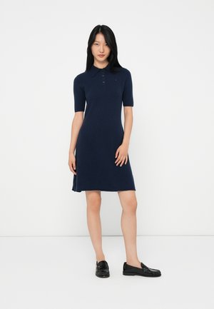 Kvinne som står mot en hvit vegg iført en marineblå kortermet polokjole og svarte loafers, poserer med den ene hånden nær låret.