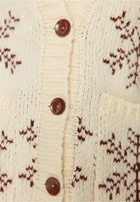 Gestrickte Strickjacke in Creme mit braunen Schneeflockenmuster, mit strukturierter Oberfläche, gerippten Kanten und vier runden, dunklen Knöpfen.
