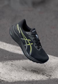 Svart och grå ASICS löparsko med gula accenter i luften ovanför asfalt, som visar den texturerade sulan och FlyteFoam-dämpningen.