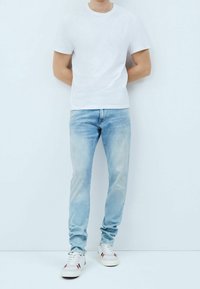 Valkoinen puuvillapaita, vaaleansiniset slim-fit farkut ja valkoiset lenkkarit, joissa on punaisia ja laivastonsinisiä yksityiskohtia, seisoo yksinkertaista taustaa vasten.