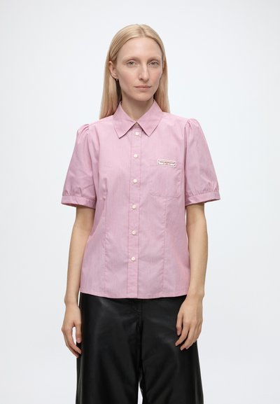 Femme aux cheveux blonds et raides porte une chemise rose à manches courtes boutonnée et un pantalon noir, debout sur un fond blanc uni.