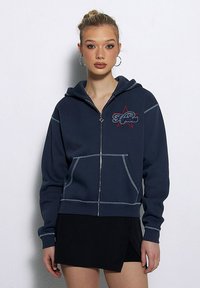 Granatowa bluza z kapturem na zamek błyskawiczny, z przednią kieszenią, kontrastowymi przeszyciami i haftowanym logo na czerwonej gwieździe. Gładki materiał, luźny krój, kaptur ze sznurkiem.