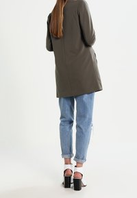Manteau long vert olive à coupe droite, associé à un jean bleu clair retroussé et des sandales blanches à talons blocs. Tissu lisse et bien ajusté.