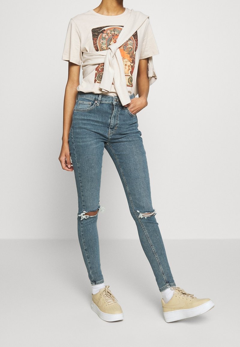 Personne portant un t-shirt graphique beige clair avec un sweat noué autour des épaules, un jean skinny bleu déchiré, des chaussettes blanches et des baskets beiges.