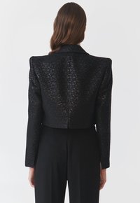 Herkömmlicher schwarzer Blazer mit strukturierten Schultern und einem klassischen Kragen. Das Material hat eine strukturierte Textur. Er weist dezente Muster auf und hat einen taillierten Schnitt.