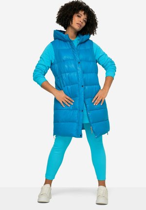 Gilet rembourré teal avec capuche, doté de boutons et de fermetures éclair, porté par-dessus un haut à manches longues assorti et des leggings. Baskets blanches.
