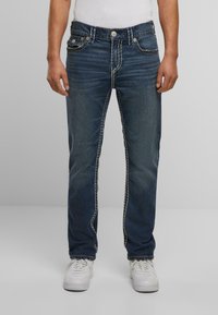 Jeans de mezclilla azul oscuro con costuras blancas en contraste, cinco bolsillos y un corte recto, combinados con zapatillas blancas.