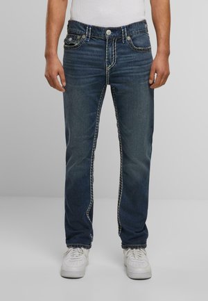 Jeans de mezclilla azul oscuro con costuras blancas en contraste, cinco bolsillos y un corte recto, combinados con zapatillas blancas.