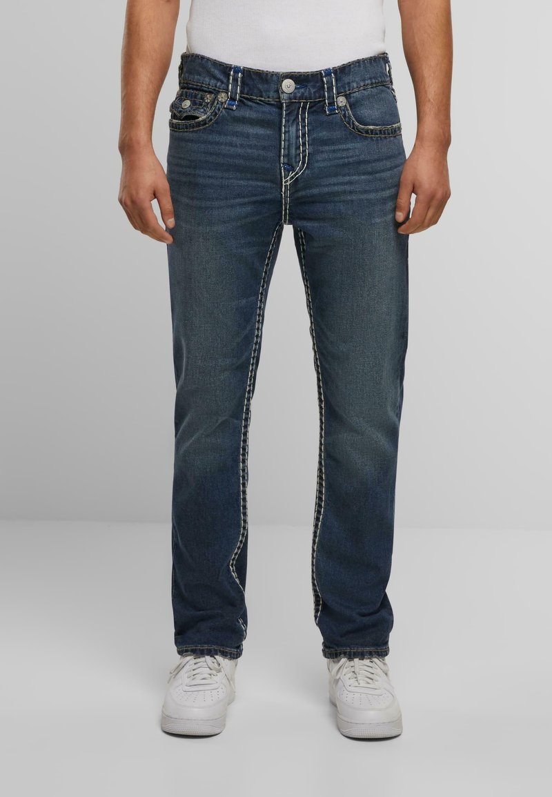 Jeans de mezclilla azul oscuro con costuras blancas en contraste, cinco bolsillos y un corte recto, combinados con zapatillas blancas.