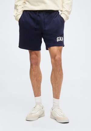 Mand iført marineblå GAP-logo shorts, hvide sokker og off-white sneakers, står med hænderne i lommerne.
