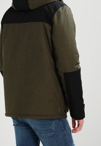 Kurtka puchowa khaki z czarnymi akcentami, posiadająca kaptur, teksturowaną tkaninę i luźny krój. Charakterystycznym elementem jest metka u dołu.