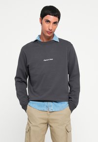 Sweatshirt gris foncé avec un col rond et du texte brodé en blanc. Superposé sur une chemise en denim bleu clair avec un col pointu.