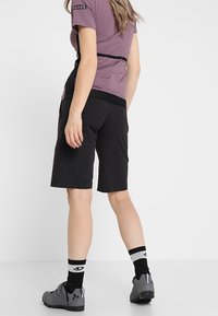 Musta pyöräilyshortsit, joissa on sileä pinta, muotoiltu malli, yhdistettynä violettiin lyhythihaiseen paita ja harmaat pyöräilykengät.