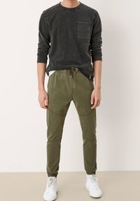 Man in een vervaagde zwarte langebloes met borstzak, olijfgroene joggingsbroek en witte hoge sneakers, staande op een neutrale vloer.