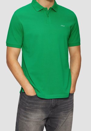 Grünes Poloshirt aus leichter Stoffqualität, mit klassischem Kragen, zwei Knöpfen und kleinem Logo auf der Brust. Kombiniert mit dunklen Jeans.