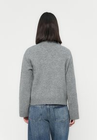 Persona con capelli scuri all'altezza delle spalle, che indossa un maglione grigio a maglia e jeans blu, di fronte a uno sfondo bianco semplice.