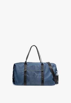 Bolsa de lona náutica de nailon azul marino con asas tejidas negras, correa ajustable, bolsillo frontal con cremallera y detalle de logotipo en relieve. Textura suave.