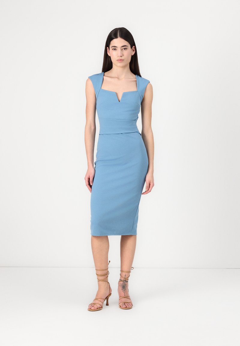 Vestido azul justo com mangas em formato de cap sleeve, decote em V e cintura acentuada. Apresenta uma textura suave e um design até ao joelho, combinado com sapatos de salto.